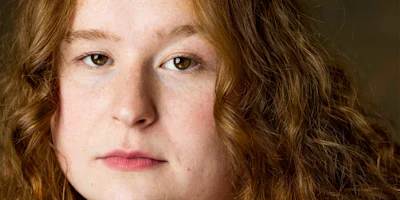 A close-up portrait of a person with long, wavy reddish-brown hair, fair skin, and a neutral facial expression.