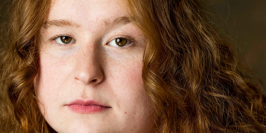 A close-up portrait of a person with long, wavy reddish-brown hair, fair skin, and a neutral facial expression.