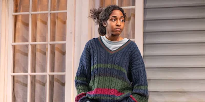 A person with curly hair wearing a striped sweater sits in front of a window and a house exterior, looking to the side with a neutral expression.