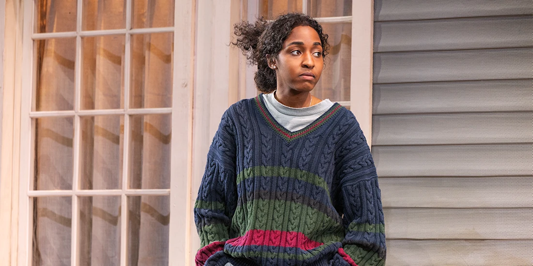 A person with curly hair wearing a striped sweater sits in front of a window and a house exterior, looking to the side with a neutral expression.