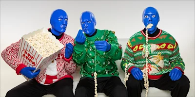 Three people with blue-painted faces wearing holiday sweaters sit on a couch. One holds a popcorn bucket, another eats popcorn, and the third has a popcorn string in their mouth.