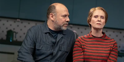 A man in a dark shirt looks at a woman in a red striped sweater who stares ahead with a serious expression; they are seated in a kitchen.