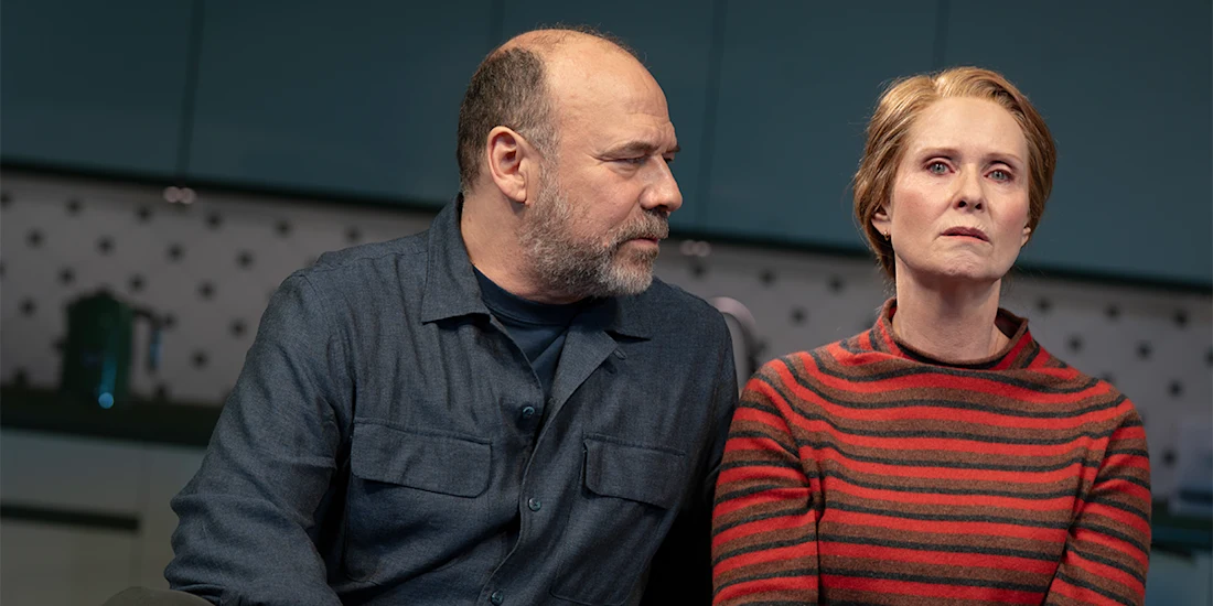 A man in a dark shirt looks at a woman in a red striped sweater who stares ahead with a serious expression; they are seated in a kitchen.