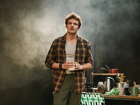A man in a plaid shirt holds a mug while standing in a kitchen setting with cooking utensils and jars on a table behind him.