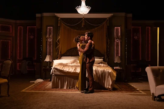 A couple embraces and kisses in the center of a lavish, dimly lit bedroom with an unmade bed and a chandelier overhead.