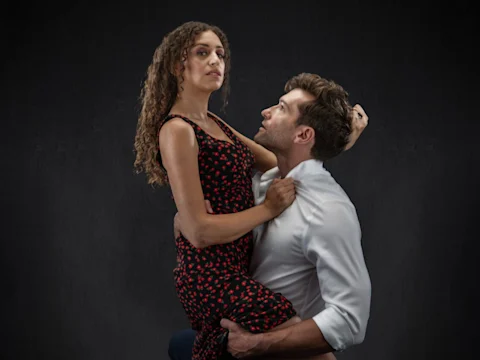 A woman in a black dress with red patterns sits on a man's knee while he holds her and looks up at her against a dark background.