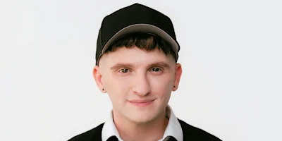 A person with short brown hair wearing a black baseball cap, black sweater, and white collared shirt, facing the camera against a plain white background.