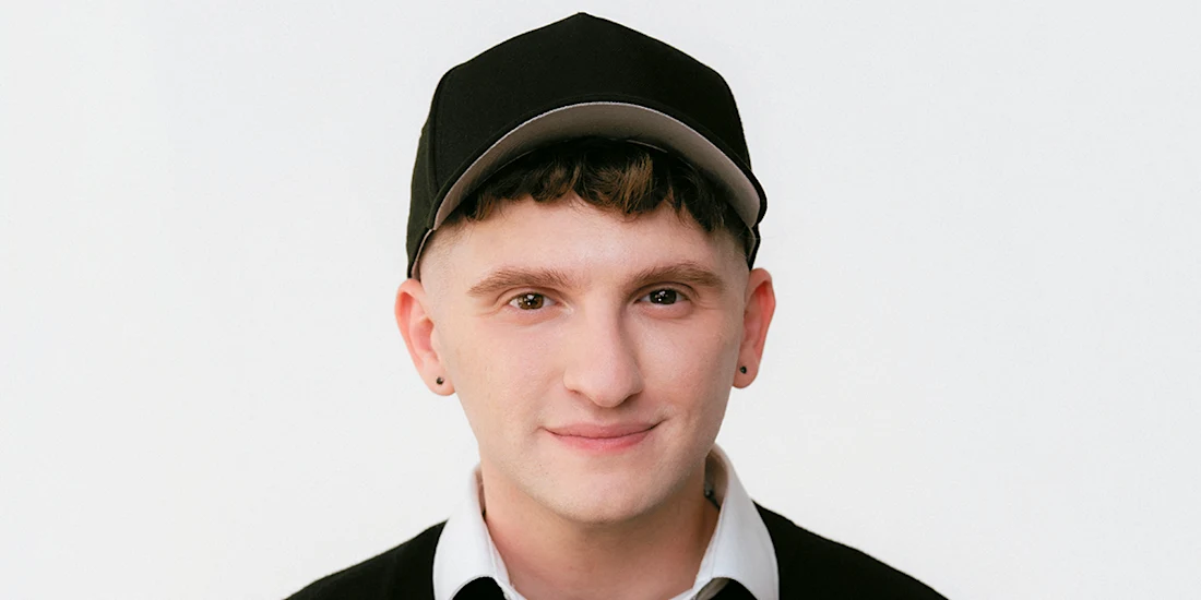 A person with short brown hair wearing a black baseball cap, black sweater, and white collared shirt, facing the camera against a plain white background.