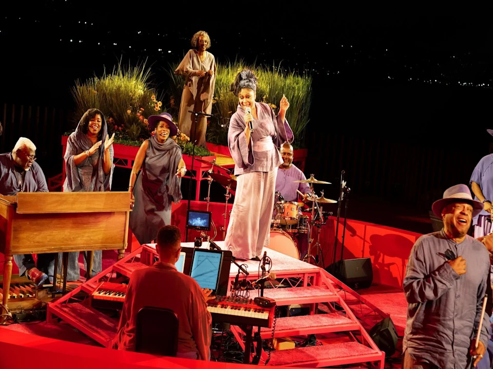 A group of musicians and singers perform energetically on a red stage with instruments and microphones, under stage lights, with plants and a night sky in the background.