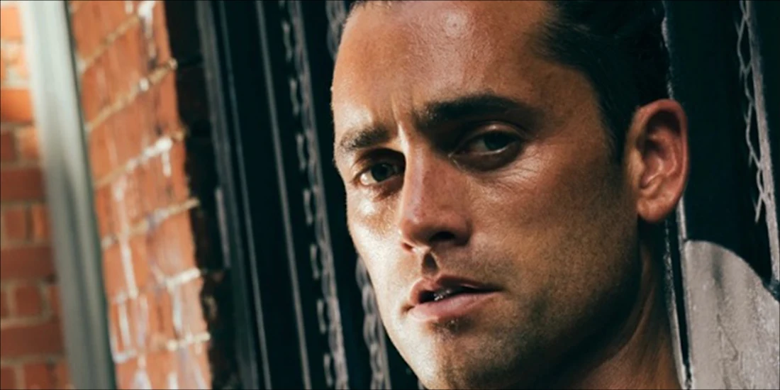 A man with slicked-back hair and stubble stands in front of a brick wall, looking directly at the camera with a neutral expression.