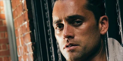 A man with slicked-back hair and stubble stands in front of a brick wall, looking directly at the camera with a neutral expression.