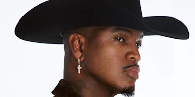 A man wearing a wide-brimmed black cowboy hat, a cross earring in his left ear, and a dark jacket, shown in profile against a plain white background.