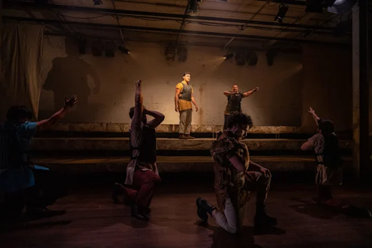 Six performers on a dimly lit stage, with two standing on elevated steps and four kneeling in dramatic poses, facing different directions.