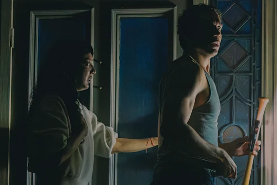 A startled woman in a white robe reaches for a man gripping a baseball bat in a dark, blue-lit hallway.