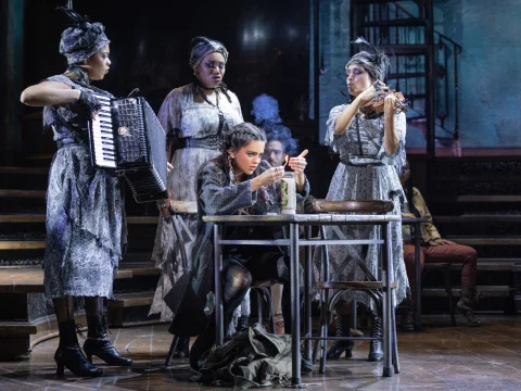 A young woman sits at a table, focusing on a document, while three performers in elaborate gray costumes play musical instruments around her on a dimly lit stage.