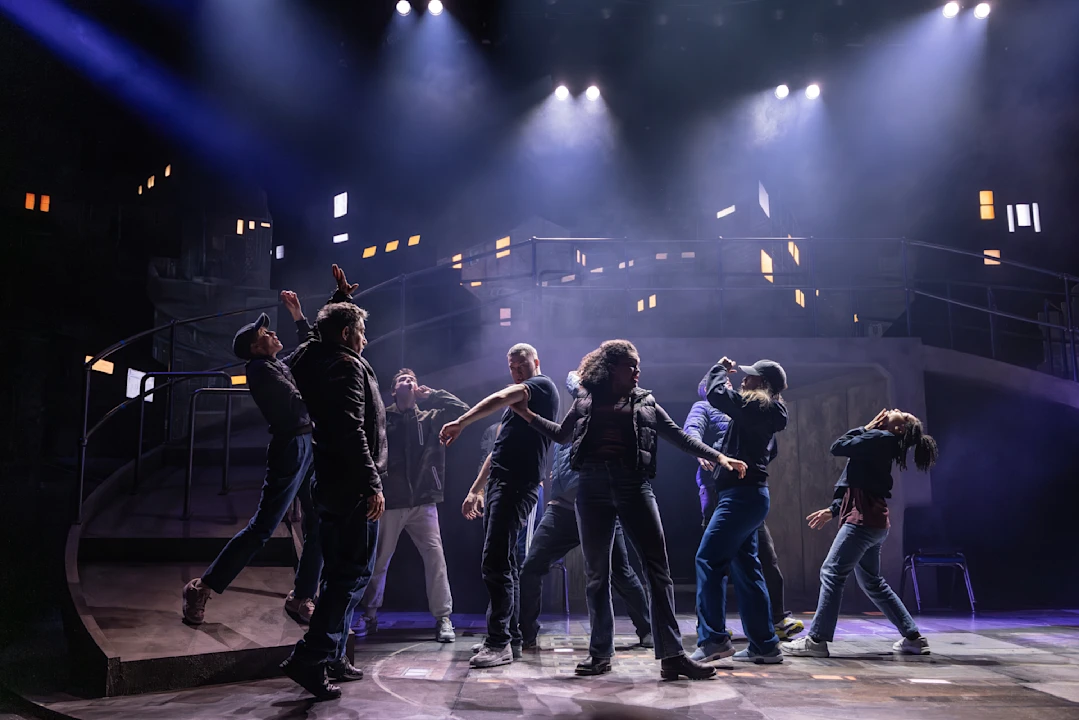 A group of actors perform a dramatic scene on a dimly lit stage with an urban set and spotlights overhead.