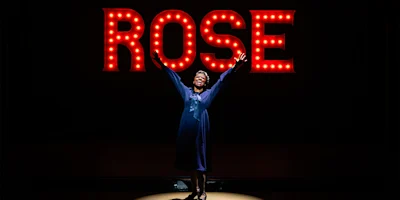 A person in a blue dress stands center stage with arms raised in front of a large illuminated sign that reads "ROSE.