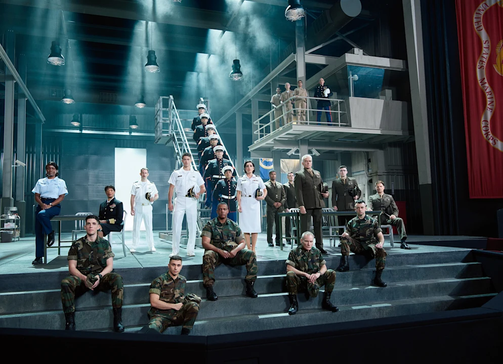 A group of U.S. military personnel in various dress and camouflage uniforms posed on and around a set of stairs in a studio environment.