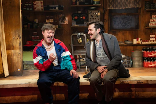 Two men sit on a wooden stage, laughing and talking. One wears a colorful windbreaker and jeans; the other wears a sweater and coat. The background is a cluttered, rustic set.