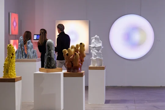 Three people view ceramic sculptures on pedestals and illuminated abstract artworks mounted on the walls in a contemporary art gallery.
