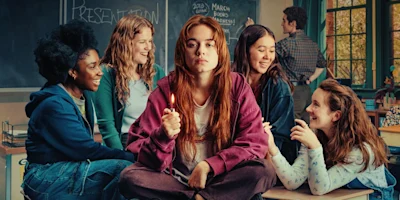 A young woman with red hair sits in the center of a classroom holding a lit lighter, surrounded by four smiling friends.