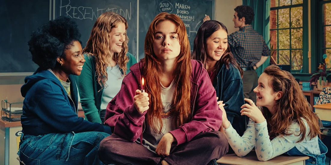 A young woman with red hair sits in the center of a classroom holding a lit lighter, surrounded by four smiling friends.