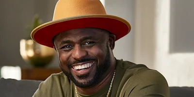 A man with a beard smiles while wearing a tan and orange wide-brimmed hat, a gold chain, and an olive green shirt, seated indoors.
