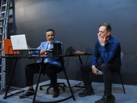 Two men are seated in an office; one on the left is speaking and working on a laptop, and the other on the right appears contemplative with his hand on his chin.