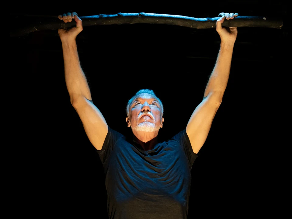 A person with a beard and mustache holds a long stick above their head with both arms, standing against a dark background.