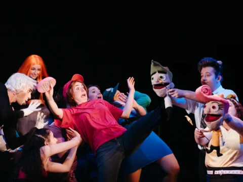 A group of people are performing an animated scene on stage with puppets. The characters are dressed in colorful costumes, and one person in a red shirt and cap is at the center, mid-action.