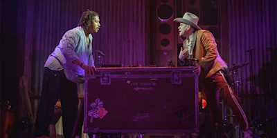 Two men stand and face each other across a large equipment case on a stage, with dramatic lighting and musical instruments in the background.