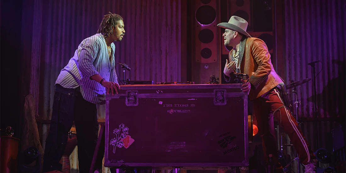Two men stand and face each other across a large equipment case on a stage, with dramatic lighting and musical instruments in the background.