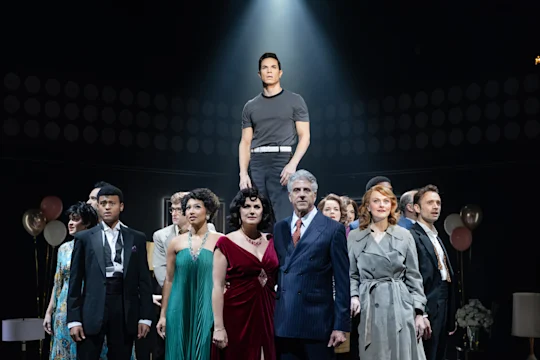 A group of people in formal and semi-formal attire stand together on stage, with one person elevated at the center under a spotlight.