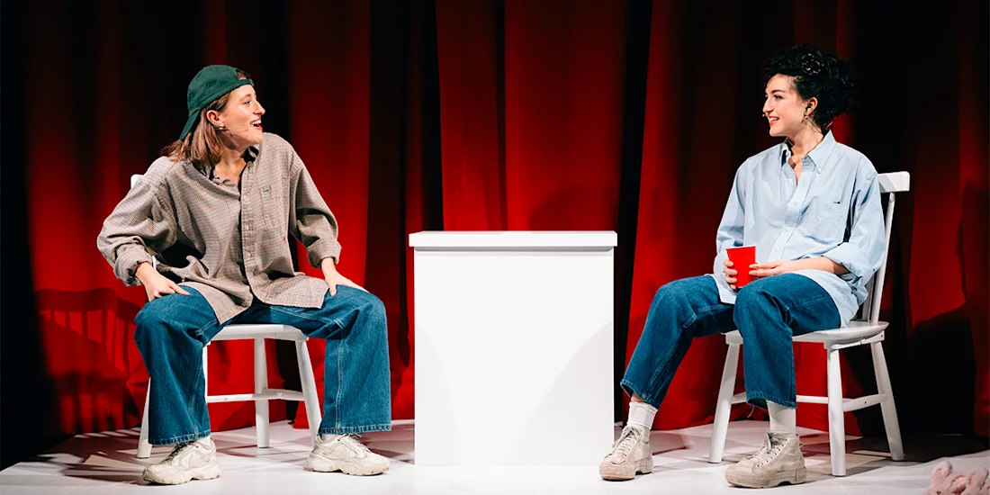 Two people sit in chairs on a stage in front of red curtains, separated by a white pedestal.