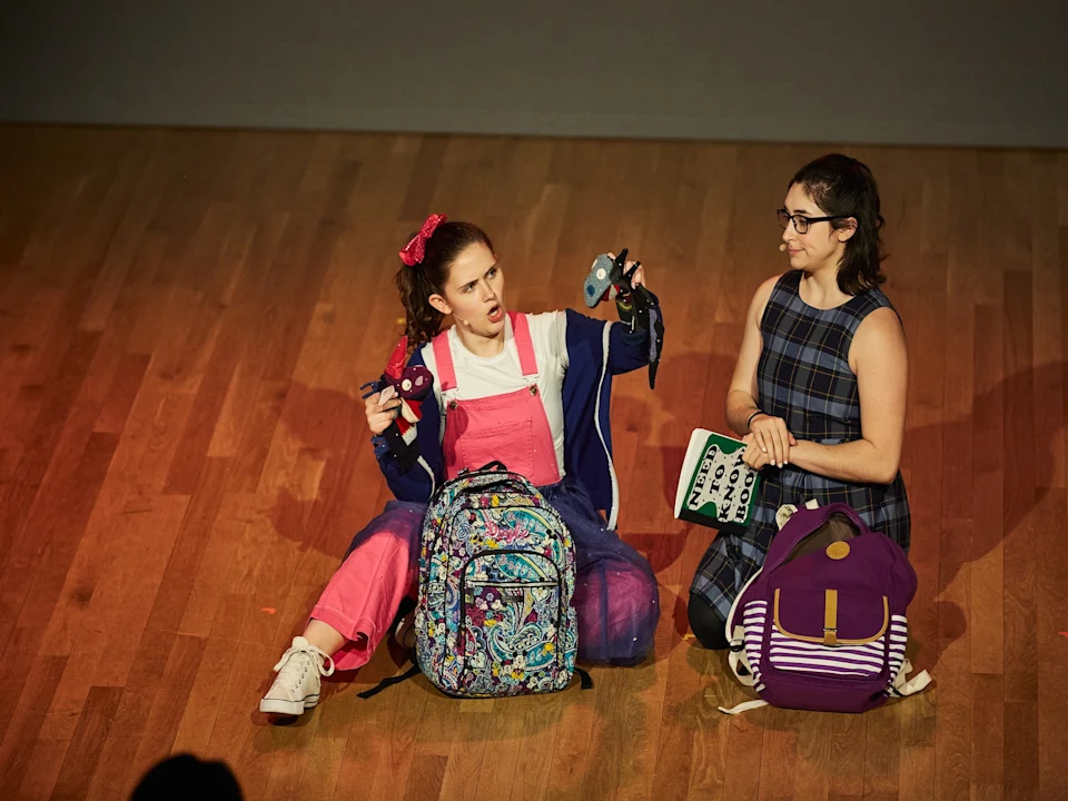 Two young women sitting on a wooden floor with backpacks; one is holding a plush toy and gesturing animatedly, while the other smiles and holds a notebook.