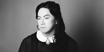 Person with long dark hair wearing a vintage-style dress with a white collar and brooch, looking away with a neutral expression against a plain background. Black and white photo.