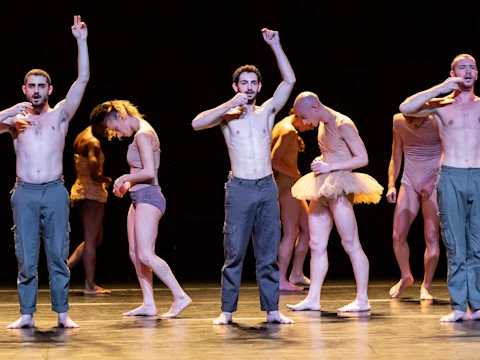 Group of six dancers in various poses on stage, some wearing partial costumes. The scene is dimly lit with a focus on the performers' expressive movements and body language.