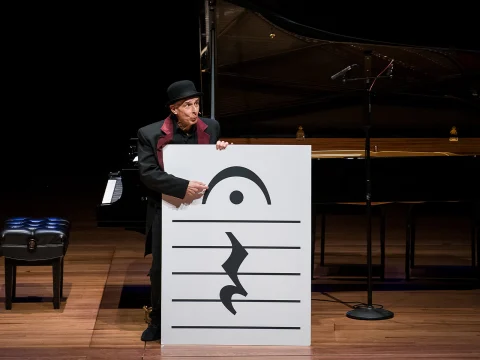 A performer in a black hat and coat holds a large sign with musical notes in front of a grand piano on a stage.