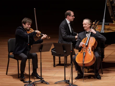 Three musicians perform on stage, featuring a violinist, a pianist, and a cellist. The violinist and cellist are seated while the pianist performs in the background.