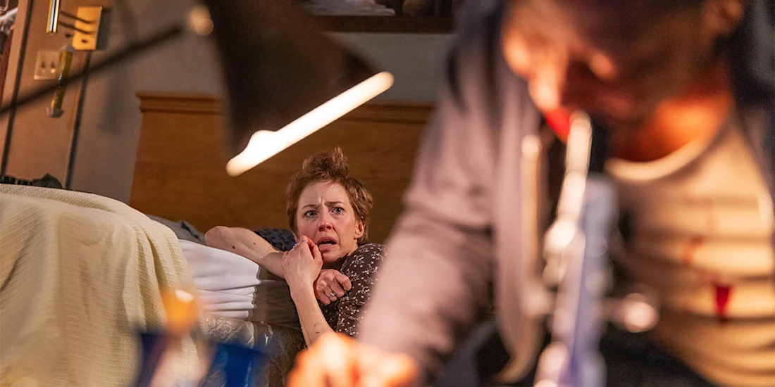 A distressed woman sits on the floor by a bed, looking fearful, while a man in the foreground is partially out of focus, leaning over a table under a lamp.