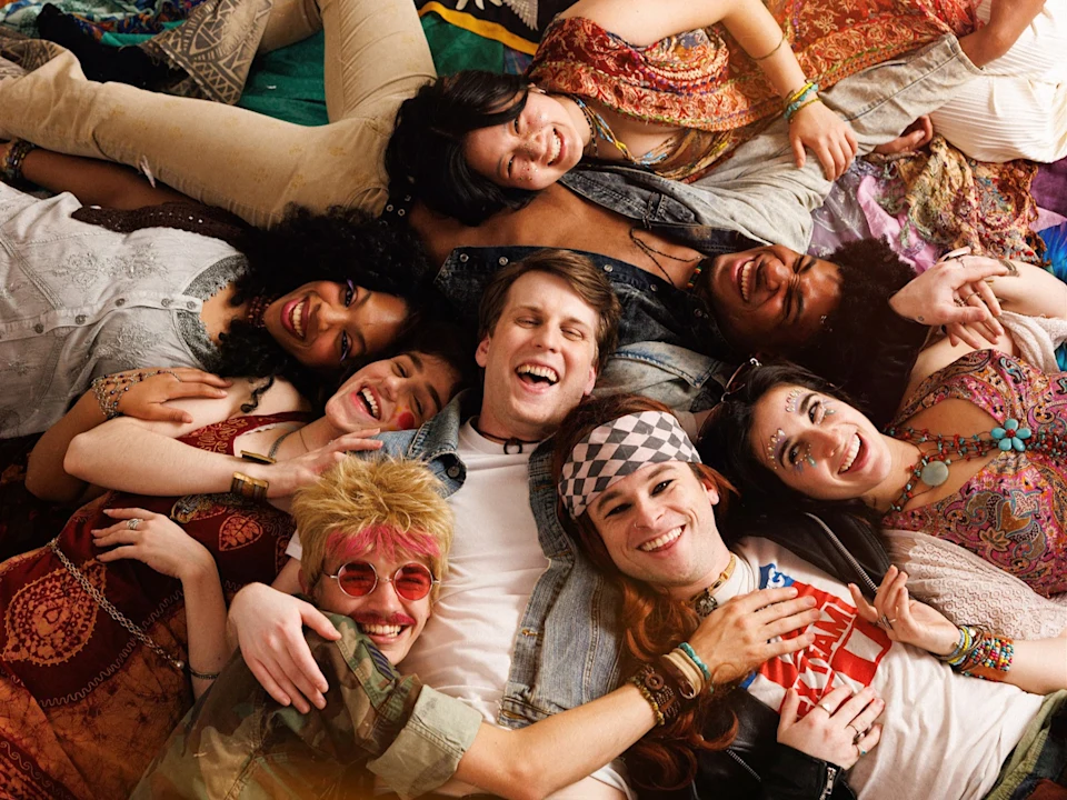 A group of eight people lie close together on the floor, smiling and laughing, dressed in colorful, bohemian-style clothing and accessories.