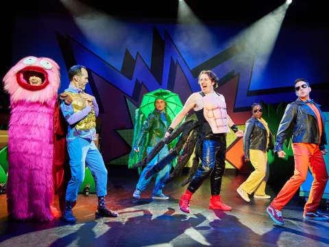 Performers in colorful, eclectic costumes are on stage, with one dressed in a pink furry suit, another with armor, and a third with a muscle top and wings. The backdrop has a vibrant, abstract design.