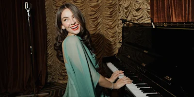 A woman in a teal dress smiles while playing a black upright piano.