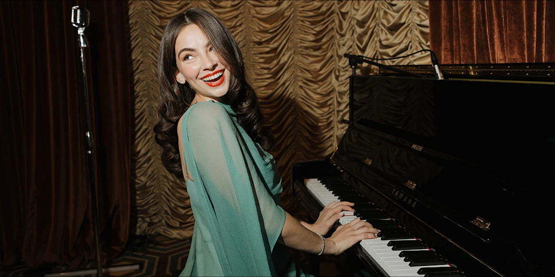 A woman in a teal dress smiles while playing a black upright piano.