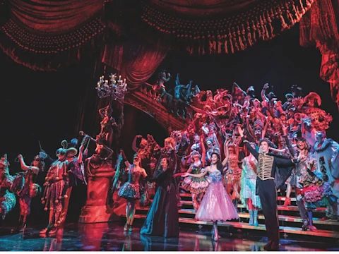 A large cast in colorful costumes performs a theatrical scene on a grand stage with dramatic lighting, elaborate drapery, and a staircase centerpiece.