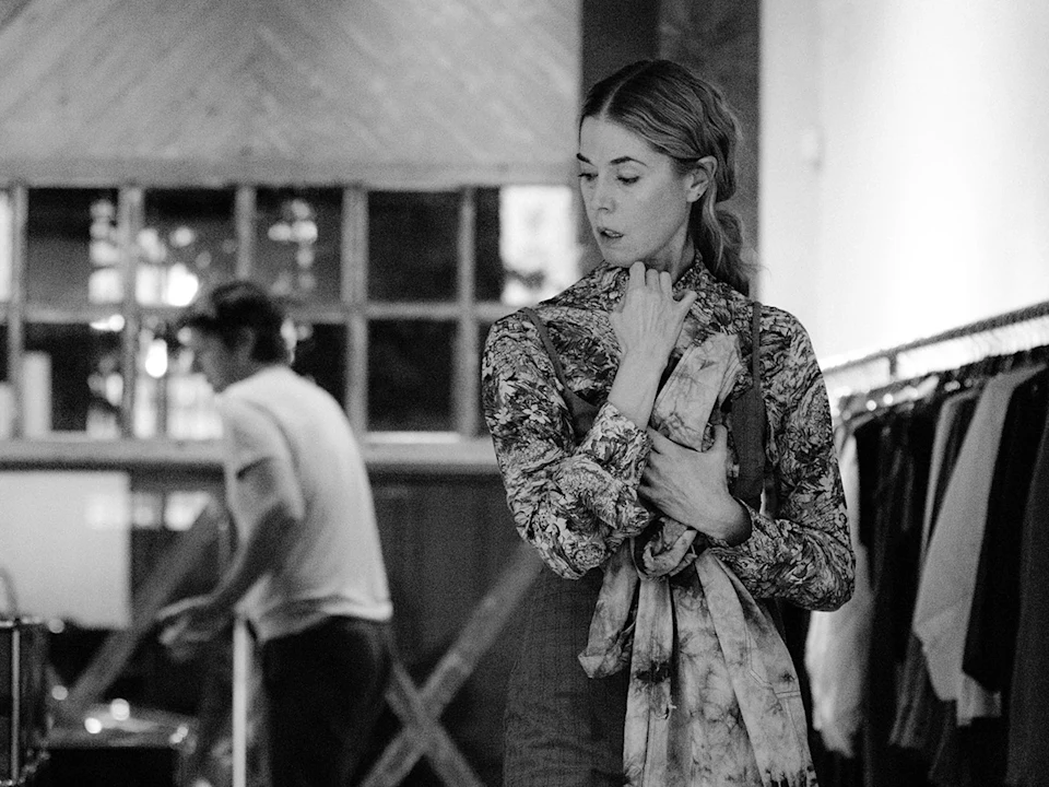 A woman stands in a clothing store holding garments, looking to the side, while a man is seen blurred in the background near racks of clothes.
