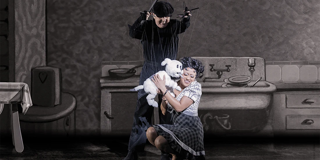 A woman dressed in vintage clothing holds a white puppet dog while a puppeteer in black manipulates strings, set against a monochrome kitchen backdrop.
