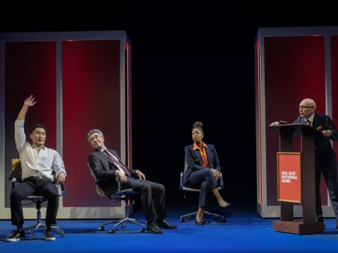 Four people in formal attire are on stage: two seated in chairs, one with hand raised, one at a lectern with "Far East National Bank," and one seated without interaction. Red panels in the background.