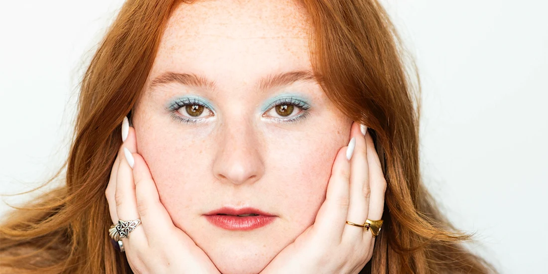 A person with long red hair rests their face in their hands, looking directly at the camera. They wear light blue eyeshadow and rings on both hands.