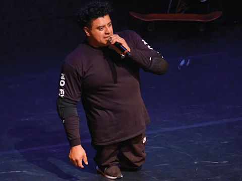 A man wearing black athletic clothing and kneepads speaks into a microphone while kneeling on stage; a skateboard is visible in the background.